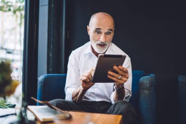 Şaşırdım, olgun kel erkek resmi kıyafetler giyiyor modern tablet kullanıyor ve kafedeki masada şaşkın şaşkın oturuyor.