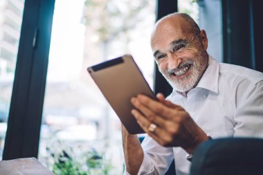 Neşeli, gülümseyen, kel, resmi kıyafetler giyen, modern tabletlere göz atan, kafedeki koltukta oturan yaşlı bir erkek.