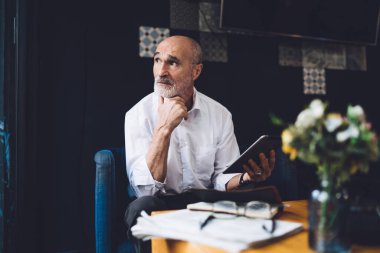 Endişeli, kel, sakallı, gri saçlı yaşlı bir adam. Gözlüklü, oturma odasının yanında otururken tablet kullanıyor.