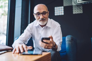 Beyaz gözlüklü, gri sakallı yaşlı erkek mavi sandalyede oturuyor ve tablette sörf yaparken akıllı telefon kullanıyor.