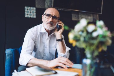 Ciddi, kel, sakallı, gözlüklü, beyaz gömlekli, mavi sandalyede oturan ve telefonla konuşurken yüzünü çeviren yaşlı bir erkek. 