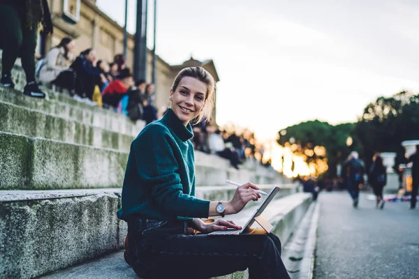 Portrait of positive hipster girl with electronic pen in hand resting at city urban area and using public internet connection for creating media sketches via application for creative artists
