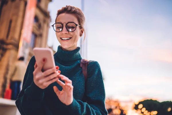 ソーシャルメディアアプリでメッセージを発信しながら 夜の明るい通りに立って笑っている眼鏡の喜びの女性学生の低角度 — ストック写真