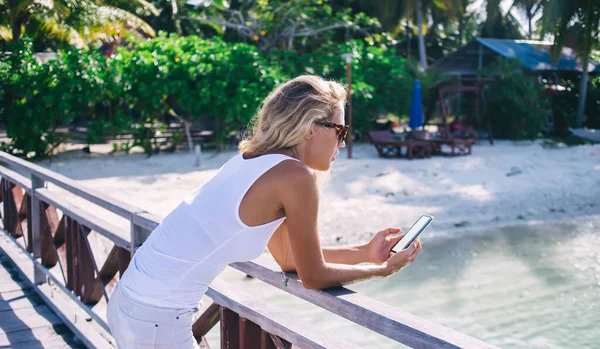 身穿白色上衣短裤 戴着太阳镜的瘦小金发女子 靠着码头栏杆 在海上滑行 白天在朦胧的海滨背景下在手机上打字 — 图库照片