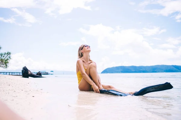 年轻美丽的金发姑娘 戴着太阳镜 身穿黄色泳衣 头戴拖鞋 坐在沙滩上 仰望高山 云彩和大海的背景 — 图库照片