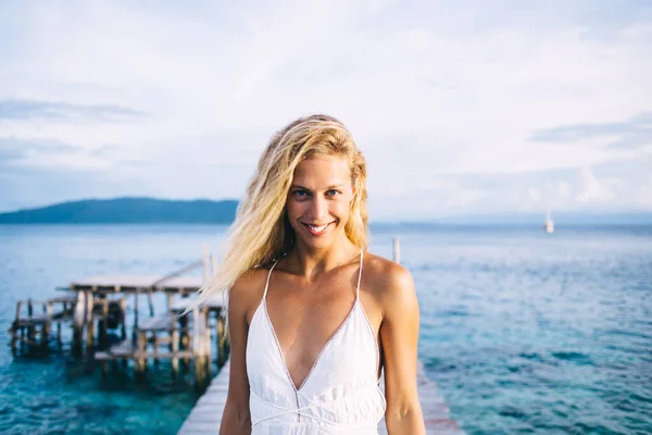 笑顔形日焼け若いですブロンド女性で白いドレス上の木製の桟橋に立って海と見てカメラで日中にぼやけた背景 — ストック写真