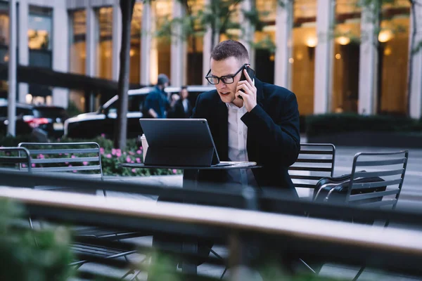 年轻英俊的经理戴着眼镜 一边认真地在智能手机上说话 一边仔细地看着平板电脑屏幕 一边坐在街边咖啡馆的桌旁 — 图库照片