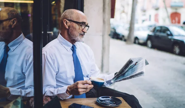 一位严肃的长发老人 身穿白衬衫 头戴咖啡杯 在靠近玻璃和道路的城市街道上看报纸 — 图库照片