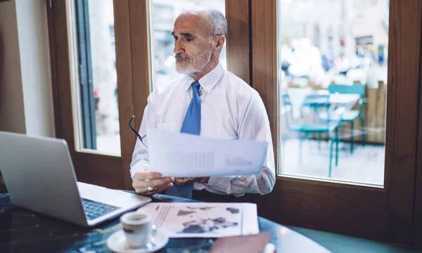 Senior bearded man sitting with paper and glasses in hands in front of laptop located on table near with cup of coffee and notebook