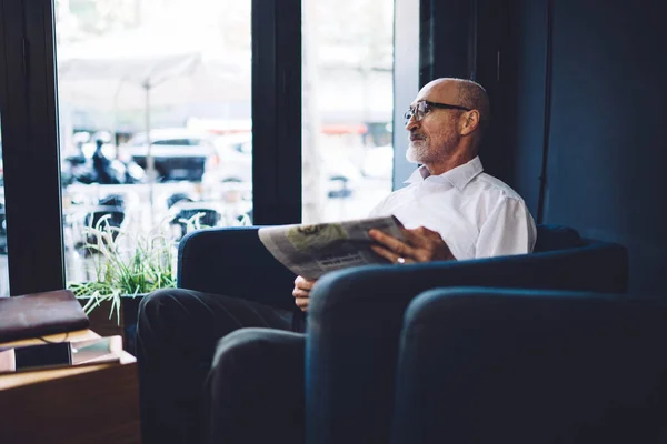 穿着白衬衫和眼镜的老秃头男子坐在咖啡店舒适的椅子上 一边看一边看报纸的侧影 — 图库照片