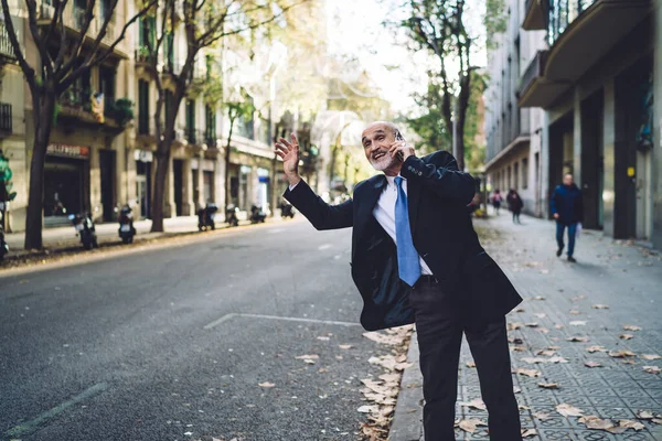 Smiling handsome old male in suit and with white beard having phone call while stopping car on roadside of street looking away on background of buildings