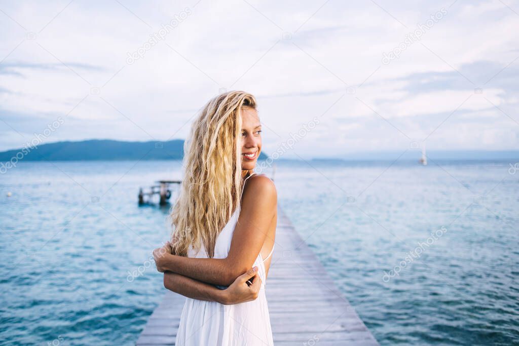 Happy bronzed young blonde woman in white dress with open back standing on wooden pier at sea and looking away