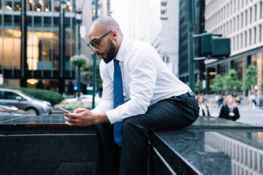 Beyaz tişörtlü, mavi kravatlı, şık, kel bir erkek çalışanın sokak merdivenlerinde ticari binaların arka planında sörf yaparken görüntüsü.