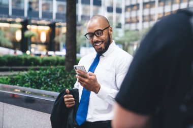 Ofis kıyafetleri ve gözlüklü, takım elbiseli neşeli siyah adam modern ofis binasının önünde dikilirken akıllı telefon kullanıyor.