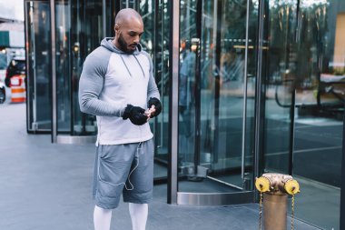 Yakışıklı, kel, sakallı, spor kıyafetli, kulaklık takan, modern ofis binasının yanındaki yangın musluğuyla caddede duran Amerikalı adam.