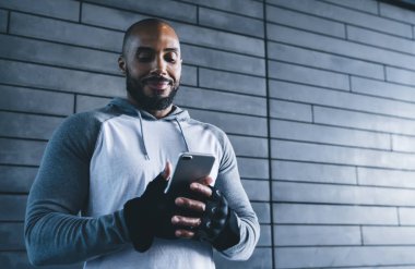 Gri kazaklı ve eldivenli atletik siyah vücutgeliştiricinin spor salonunda gri tuğladan duvarlı spor yaparken cep telefonuna bakması.