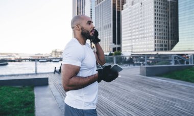 Spor giyimli, kel, sakallı, spor kıyafetli, akıllı bir erkek, şehir merkezindeki ahşap iskelede kulaklık takıyor.