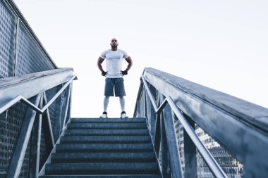 Spor giyimli, kel, sakallı, spor kıyafetli bir adam merdivenlerin tepesinde el ele tutuşup, spor yaparken kalçalarını tutuyor.