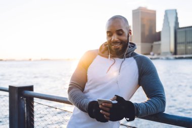 Neşeli Afro-Amerikan sportif sörf arabalı adam sabah güneşinde setin kenarındaki çitin yanında duran kulaklıkları takıyor.