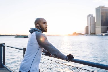 Kulaklıklı ve spor elbiseli dikkatli etnik erkeğin nehir kenarında durup düşünceli bakışlarını başka yöne çevirmesi.