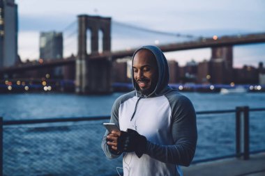 Keyifli, kaslı, spor kıyafetli, akıllı telefon kullanan ve akşamları bulanık arka planda, Megapolis 'teki rıhtımda dinlenirken müzik dinleyen kaslı bir adam.