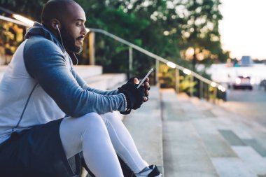Taş merdivenlerde oturan ve akıllı telefondan mesajlaşırken kulaklıkla müzik dinleyen Afrikalı Amerikalı genç sporcunun yan görüntüsü