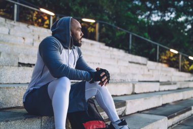 Genç Afro-Amerikan atletik erkek sırt çantasının yanında oturuyor ve kulaklıkla müzik dinliyor.