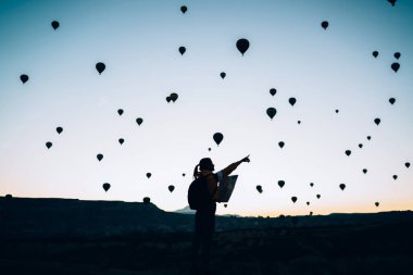 Sırt çantalı, kayalık arazide elinde haritayla duran ve etrafta yüzen hava balonlarını gösteren kimliği belirsiz bir kadının arka planı.