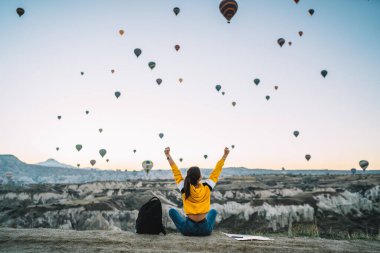 Huzurlu bir kadının kayalık arazide uçurumun kenarına oturmuş gün batımında hava balonlarına bakarken.