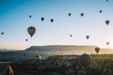 Hava manzaralı renkli çizgili sıcak hava balonları şafak vakti açık gökyüzü ve yandan gelen güneş ışığıyla dağlık bölgelerde birbirleriyle yarışıyor.