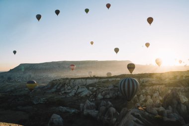Hava manzaralı arkadaki sıcak hava balonları şafak vakti dağlık arazide birbirleriyle yarışıyorlar ve açık gökyüzüyle güneş ufukta yükseliyor.