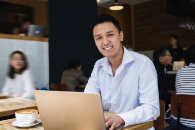 Yetişkin, yakışıklı, gülümseyen Asyalı, resmi kıyafetler giymiş, laptopta gezinen ve kahve dükkanındaki kameraya bakan bir erkek.