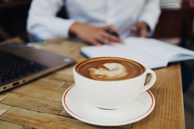 Yukarıdaki büyük beyaz kupadan kapuçino kapuçinosu üzerinde, kalbi köpüklü, ahşap masa üzerinde çalışan ofis çalışanına servis edilir.