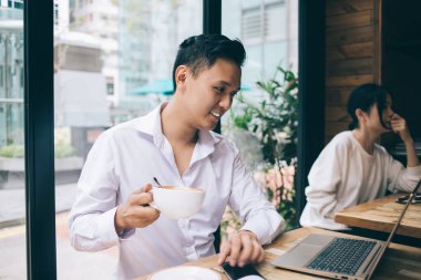 Beyaz gömlekli Asyalı yakışıklı genç adam laptopta çalışıyor ve kafeteryada pencerelere gülümsüyor.
