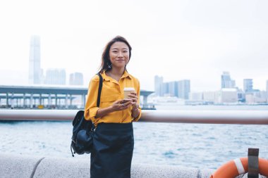 Günlük kıyafetlerle gülümseyen Asyalı güzel bir kadın ve elinde kahve fincanı ve akıllı telefonla rüzgarlı bir günde Hong Kong rıhtımında dikilirken siyah sırt çantasıyla.