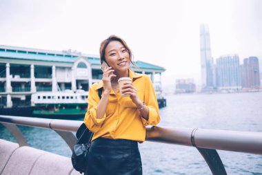 Çekici, neşeli Asyalı kadın, sarı gömlekli, siyah deri etekli, elinde kahve tutan Hong Kong 'daki feribot iskelesinde akıllı telefondan konuşmak için.