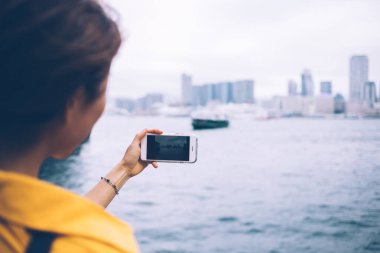 Sarı bluz giyen kadın turistin, çirkin bir günde feribot ve modern gökdelenlerle güzel şehir körfezinin yatay fotoğrafını çekerken görüntüsü.