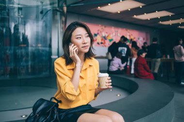 Güzel, sıska, genç Asyalı bir kadının dijital akıllı telefonuyla iletişim kurmasına ve aramasına sevindim. Bir bardak içkiyle birlikte otururken ve modern tasarımla aynı odaya bakarken.