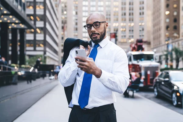 Confident focused African American elegant clothed male with eyewear and jacket over shoulder chatting on mobile phone on street in downtown