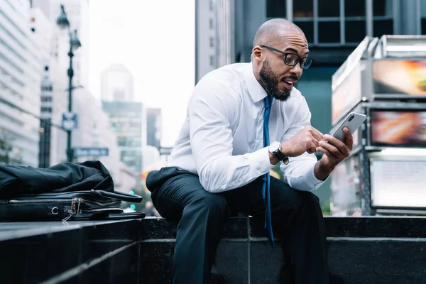 Excited formal African American male employee reading message on phone with amazement while sitting on street stairs with leather bag on background of blurred commercial building