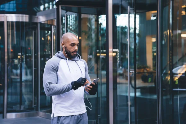 在训练期间 英俊的黑人运动员站在靠近大楼玻璃入口的街上 一边用耳机一边听音乐一边浏览社交媒体 — 图库照片