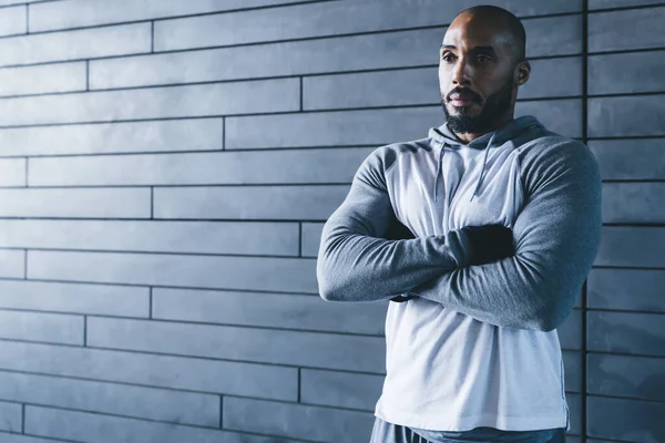 Yakışıklı, kaslı, Afro-Amerikan kel, sakallı, spor giyimli, kollarını çapraz bağlamış yetişkin erkek kapalı alanda gri arka plana bakıyor.