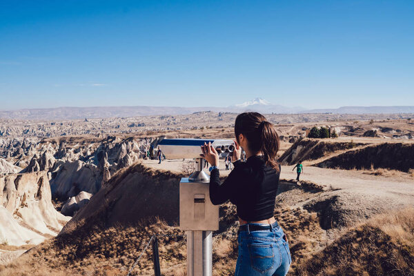 Back view of slim young lady standing near binoculars on observation platform and looking at rocky terrain against cloudless blue sky in nature