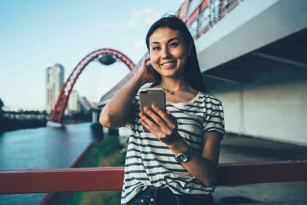 Half length portrait of cheerful Asian female blogger with cellular device looking at camera and waiting text reference at own public website with lifehacks for teenagers, generation Y person