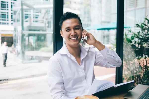 自信を持って成功した幸せなアジア人男性白いシャツのテーブルに座ってパノラマのガラス窓の近くにあり スマートフォンで会話をして — ストック写真