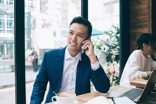 Молодий Азіатський Бізнесмен Сидить Столом Ноутбуком Посміхається Розмовляючи Смартфону Озираючись — стокове фото