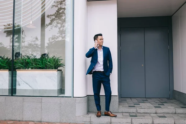 白い壁 ドアやガラスのフェンスの近くに立っているビジネススーツのアジアの笑顔の若い男は スマートフォンで話していると街の通りを離れて見て — ストック写真