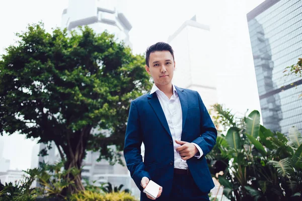 街の木々や茂みの前に立っている厳格な青いスーツときちんとした白いシャツの若いアジアの起業家が手にスマートフォンを持ち カメラを見て — ストック写真
