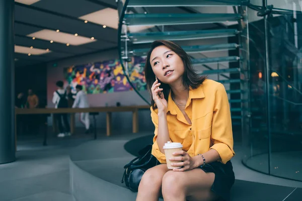 身穿休闲装的时髦亚洲女性一边拿着咖啡杯一边用手机说话 坐在现代走廊里 一边望着远方 — 图库照片