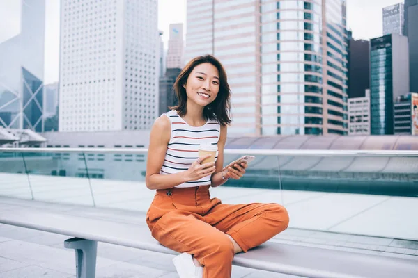 Portrait of cheerful hipster girl with cellular gadget in hand smiling at camera while resting at urban setting in metropolis, positive Asian female blogger with mobile phone and coffee to go